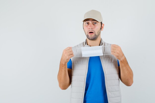 Junger Mann, der medizinische Maske im T-Shirt, in der Jacke hält und verwirrt, Vorderansicht schaut.