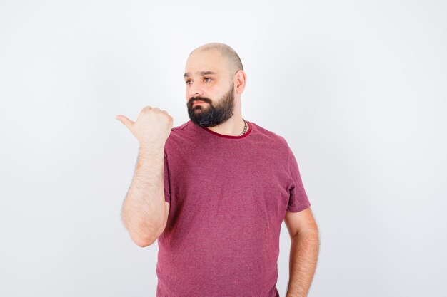 Junger Mann, der mit Daumen im T-Shirt auf die rechte Seite zeigt und zögerlich aussieht, Vorderansicht.