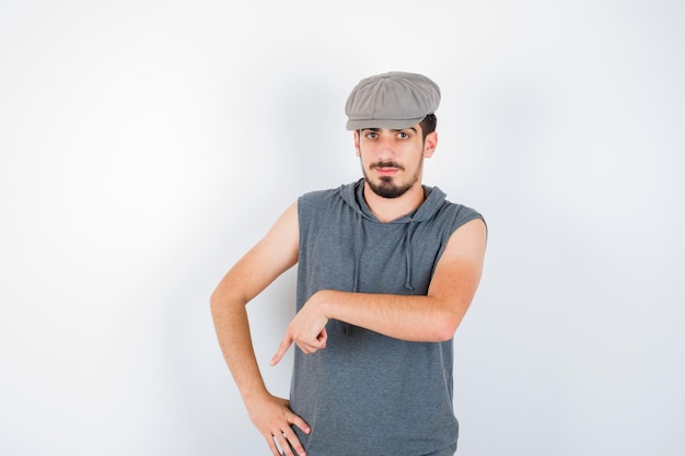Junger Mann, der mit dem Zeigefinger in grauem T-Shirt und Mütze nach unten zeigt und ernst aussieht