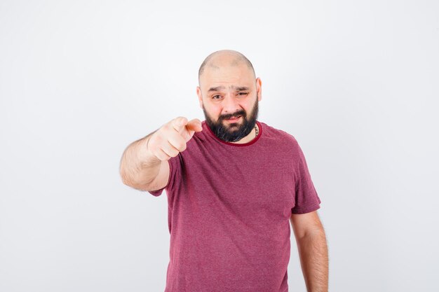 Junger Mann, der mit dem Zeigefinger in rosa T-Shirt auf die Kamera zeigt und ernst aussieht, Vorderansicht.