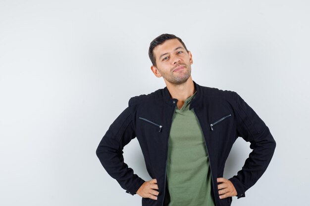 Junger Mann, der mit den Händen auf der Taille im T-Shirt, in der Jacke steht und froh schaut, Vorderansicht.