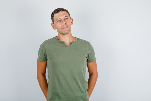 Junger Mann, der mit den Händen in den Gesäßtaschen im armeegrünen T-Shirt steht und stilvoll aussieht. Vorderansicht.