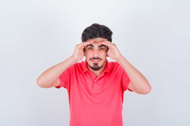 Junger Mann, der mit den Händen über dem Kopf in rosa T-Shirt schaut und lustig aussieht. Vorderansicht.
