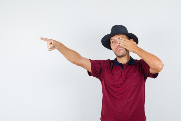 Junger Mann, der mit der Hand auf dem Auge im T-Shirt, der Vorderansicht des Hutes weg zeigt.