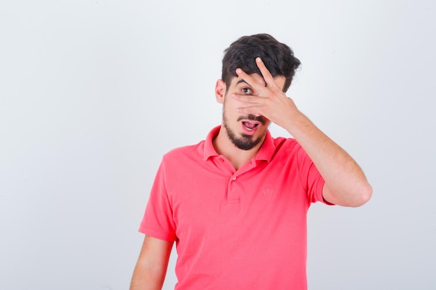 Junger Mann, der mit einem Auge zwischen den Fingern im T-Shirt herausschaut und süß aussieht, Vorderansicht.