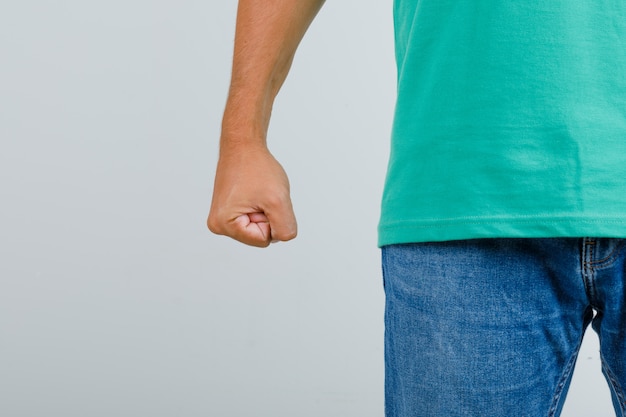 Junger Mann, der mit geballter Faust in grünem T-Shirt, Jeans steht und wütend schaut, Vorderansicht.