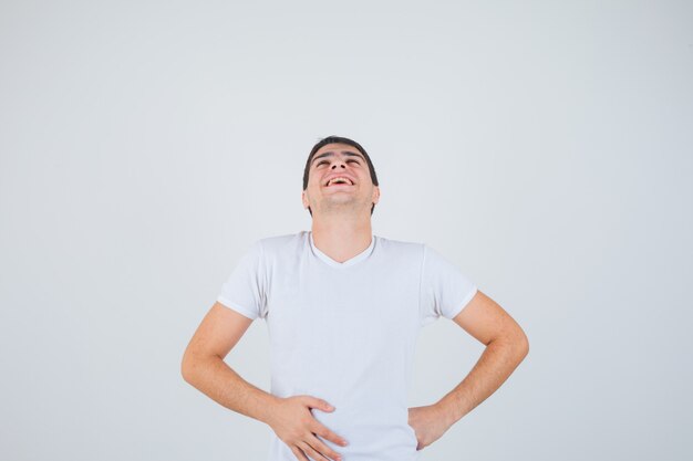 Junger Mann, der mit Händen auf Taille im T-Shirt aufwirft und freudige Vorderansicht schaut.