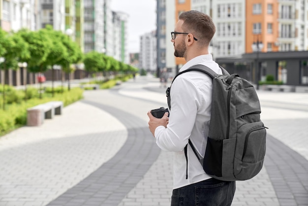 Junger Mann, der mit Rucksack und Kaffeetasse in der Stadt spazieren geht