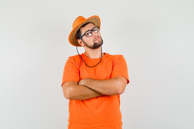 Junger Mann, der mit verschränkten Armen in orangefarbenem T-Shirt, Hut steht und nachdenklich aussieht, Vorderansicht.