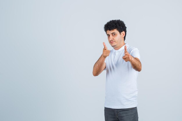 Junger Mann, der mit Zeigefingern in weißem T-Shirt und Jeans auf die Kamera zeigt und ernst aussieht, Vorderansicht.