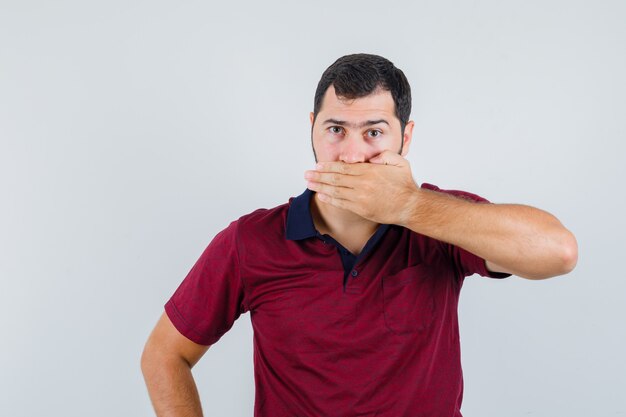 Junger Mann, der Mund mit Hand im roten T-Shirt bedeckt und ruhige Vorderansicht schaut.