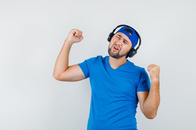 Junger Mann, der Musik genießt, während Siegergeste in blauem T-Shirt und Mütze zeigt