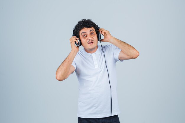 Junger Mann, der Musik mit Kopfhörern im weißen T-Shirt genießt und glücklich schaut, Vorderansicht.
