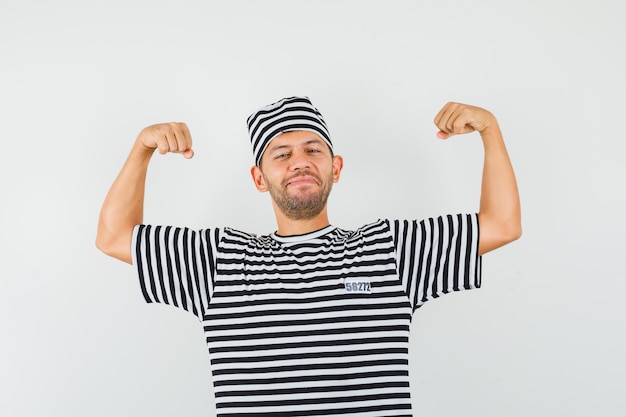 Junger Mann, der Muskeln der Arme im gestreiften T-Shirt Hut zeigt und zuversichtlich schaut