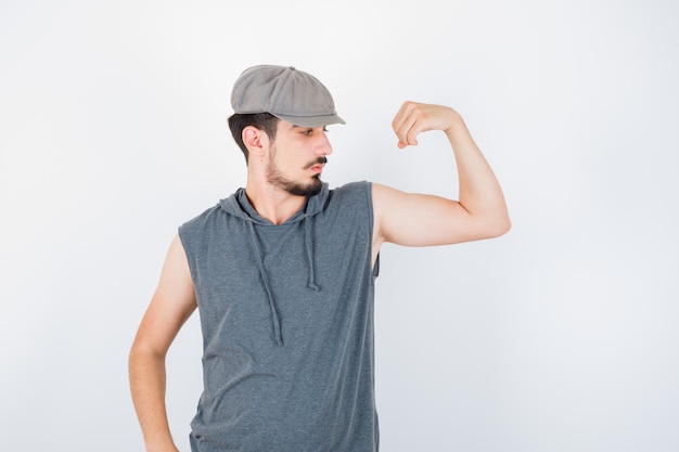 Junger Mann, der Muskeln in grauem T-Shirt und Mütze zeigt und ernst aussieht
