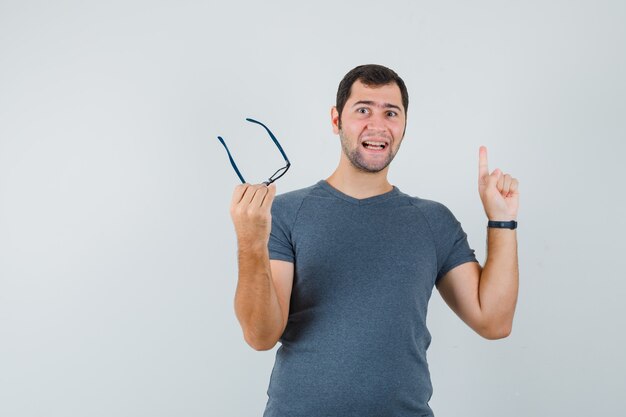 Junger Mann, der nach oben zeigt, hält Brille im grauen T-Shirt und schaut fröhlich