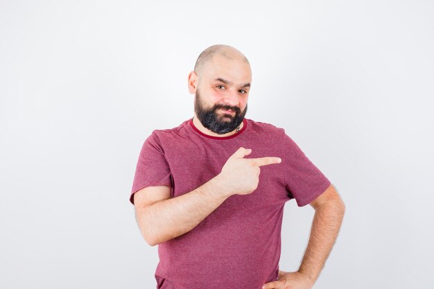 Junger Mann, der nach rechts zeigt, während er die Hand in rosa T-Shirt an der Taille hält und optimistisch aussieht, Vorderansicht.