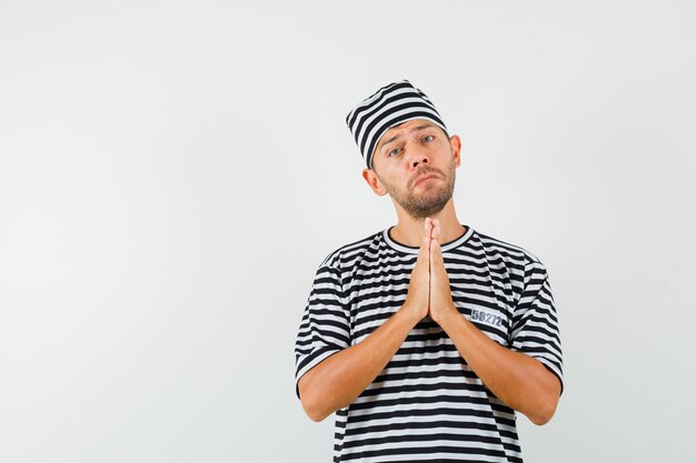 Junger Mann, der namaste Geste im gestreiften T-Shirt Hut zeigt und hilflos aussieht