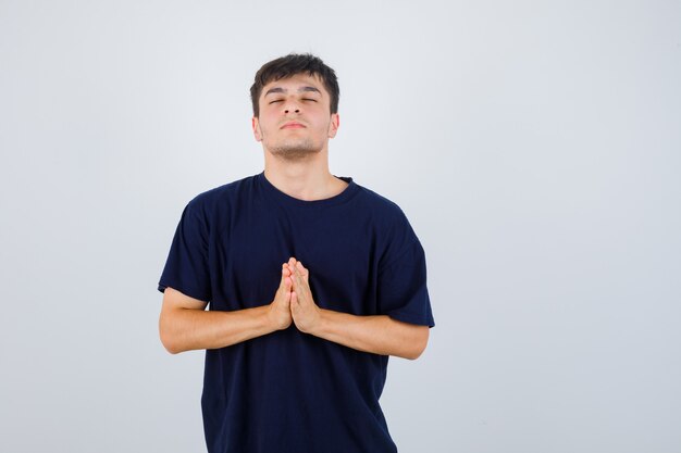 Junger Mann, der namaste Geste im schwarzen T-Shirt zeigt und hoffnungsvoll aussieht. Vorderansicht.