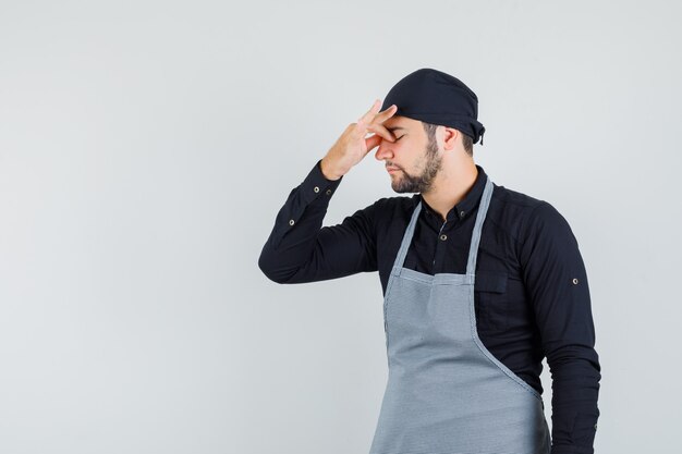 Junger Mann, der Nase und Augen in Hemd, Schürze reibt und erschöpft aussieht. Vorderansicht.