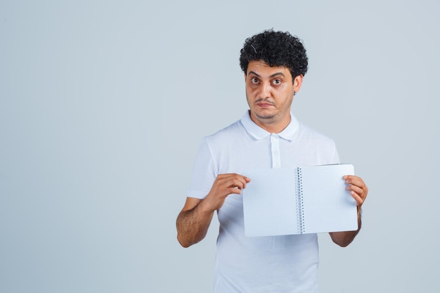 Junger Mann, der Notizbuch im weißen T-Shirt und in den Jeans zeigt und ernste, Vorderansicht schaut.