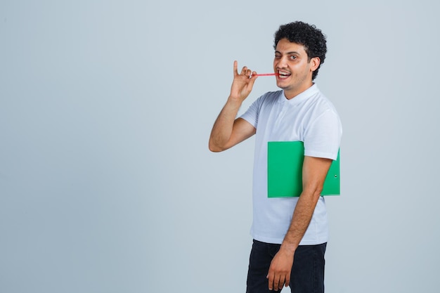 Junger Mann, der Notizbuch und Stift hält, Stift beißt, während er in die Kamera in weißem T-Shirt und Jeans schaut und glücklich aussieht. Vorderansicht.