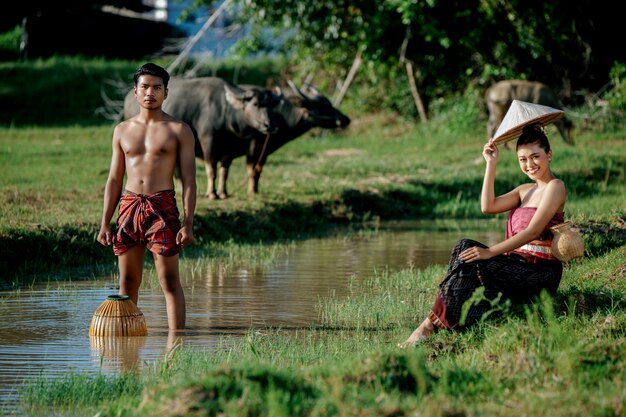 Junger Mann, der oben ohne steht und Bambus-Fangfalle hält, um Fisch zum Kochen mit einer schönen Frau zu fangen, die in der Nähe des Sumpfes sitzt?