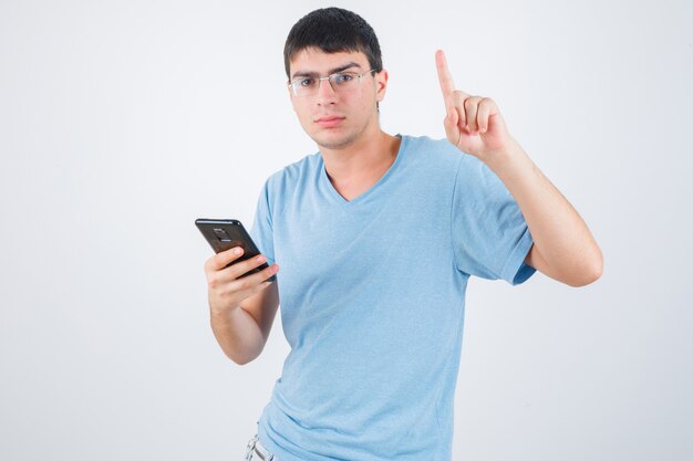 Junger Mann, der oben zeigt, während Telefon in T-Shirt hält und selbstbewusst, Vorderansicht schaut.