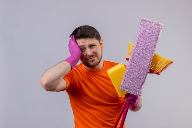 Junger Mann, der orange T-Shirt und Gummihandschuhe hält, die Reinigungswerkzeuge halten, die müde und gelangweilt mit traurigem Ausdruck auf Gesicht stehen, das über weißer Wand steht