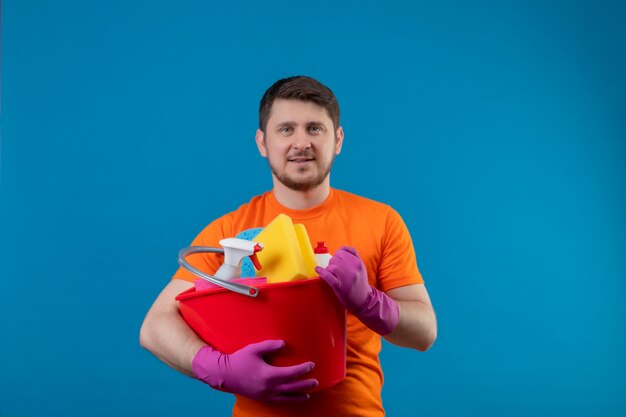 Junger Mann, der orange T-Shirt und Gummihandschuhe hält Eimer mit Reinigungswerkzeugen hält