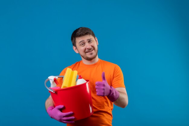 Junger Mann, der orange T-Shirt und Gummihandschuhe hält Eimer mit Reinigungswerkzeugen hält