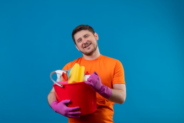 Junger Mann, der orange T-Shirt und Gummihandschuhe hält Eimer mit Reinigungswerkzeugen hält