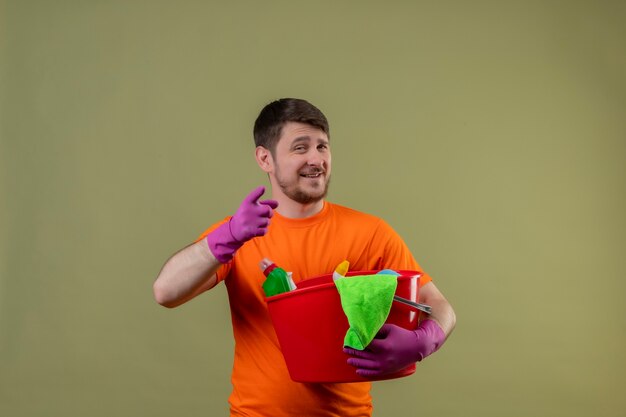 Junger Mann, der orange T-Shirt und Gummihandschuhe hält Eimer mit Reinigungswerkzeugen hält