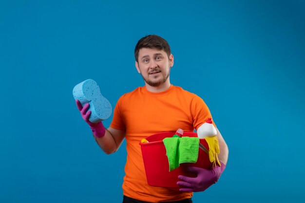 Junger Mann, der orange T-Shirt und Gummihandschuhe hält Eimer mit Reinigungswerkzeugen hält