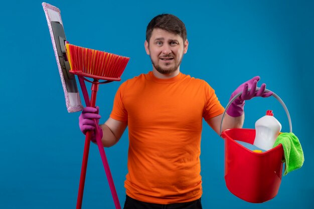Junger Mann, der orange T-Shirt und Gummihandschuhe hält Eimer mit Reinigungswerkzeugen hält