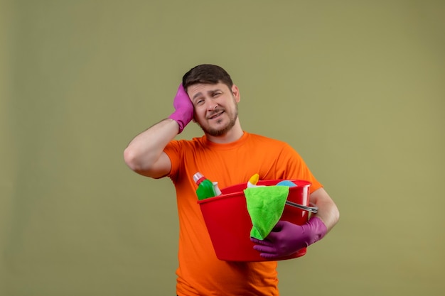 Junger Mann, der orange T-Shirt und Gummihandschuhe hält Eimer mit Reinigungswerkzeugen hält