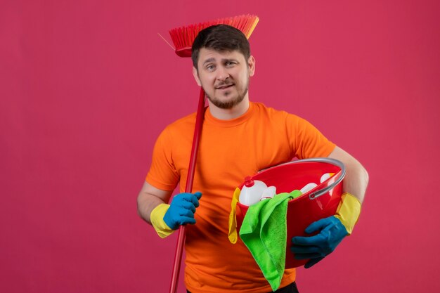 Junger Mann, der orange T-Shirt und Gummihandschuhe hält Eimer mit Reinigungswerkzeugen und Mopp hält