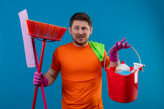 Junger Mann, der orange T-Shirt und Gummihandschuhe hält Eimer mit Reinigungswerkzeugen und Mops hält