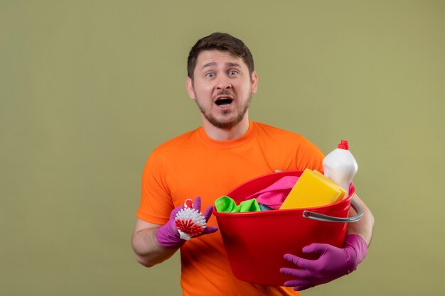 Junger Mann, der orange T-Shirt und Gummihandschuhe hält Eimer mit Reinigungswerkzeugen und Schrubbbürste hält