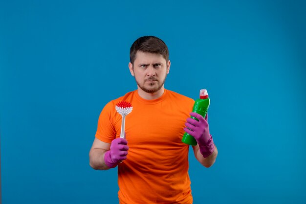Junger Mann, der orange T-Shirt und Gummihandschuhe hält Flasche mit Reinigungsmitteln und Schrubbbürste hält