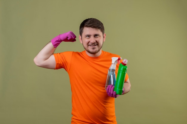 Junger Mann, der orange T-Shirt und Gummihandschuhe Reinigungsmittel trägt, lächelt fröhlich positiv und glücklich zeigt Bizeps bereit zu reinigen, über grüner Wand stehend