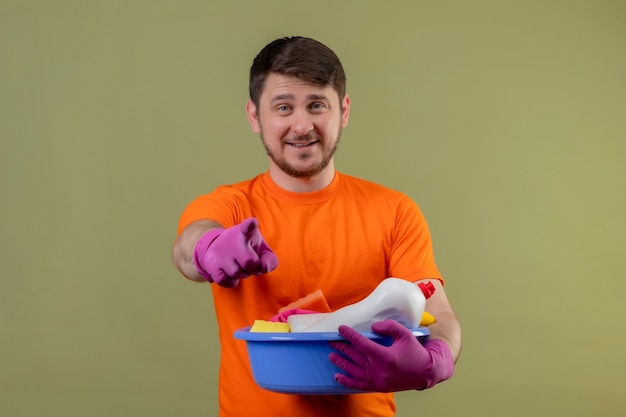 Junger Mann, der orange T-Shirt und Gummihandschuhe trägt, die Becken mit Reinigungswerkzeugen halten