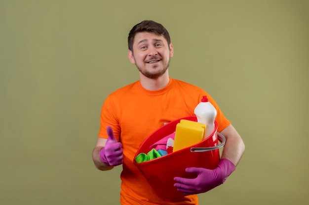 Junger Mann, der orange T-Shirt und Gummihandschuhe trägt, die Eimer mit Reinigungswerkzeugen halten, die fröhlich positiv und glücklich lächelnd Daumen hoch stehend über grüner Wand 2 lächeln