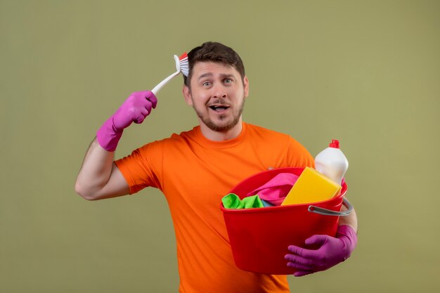 Junger Mann, der orange T-Shirt und Gummihandschuhe trägt, die Eimer mit Reinigungswerkzeugen halten, die seinen Kopf mit der Bürste kratzen