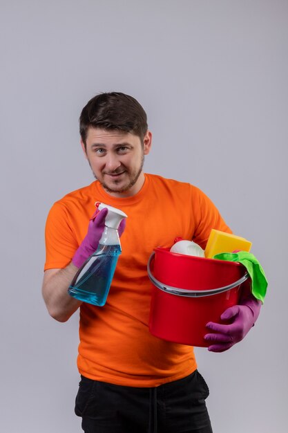 Junger Mann, der orange T-Shirt und Gummihandschuhe trägt, die Eimer mit Reinigungswerkzeugen und Reinigungsspray halten