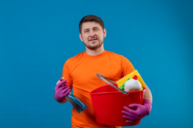 Junger Mann, der orange T-Shirt und Gummihandschuhe trägt, die Eimer mit Reinigungswerkzeugen und Reinigungsspray halten