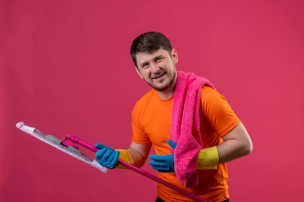 Junger Mann, der orange T-Shirt und Gummihandschuhe trägt, die Mopp und Handtuch halten, die fröhlich glücklich und positiv lächelnd über rosa Wand lächeln