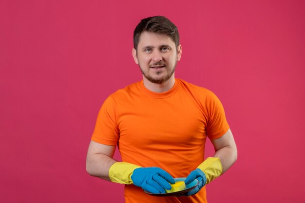 Junger Mann, der orange T-Shirt und Gummihandschuhe trägt, die Platte und Schwamm halten, die glücklich und positiv lächelnd über rosa Wand lächeln