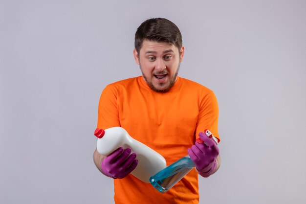 Junger Mann, der orange T-Shirt und Gummihandschuhe trägt, die Reinigungsmittel halten, die sie betrachten