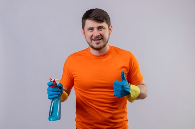 Junger Mann, der orange T-Shirt und Gummihandschuhe trägt, die Reinigungsspray lächelnd halten